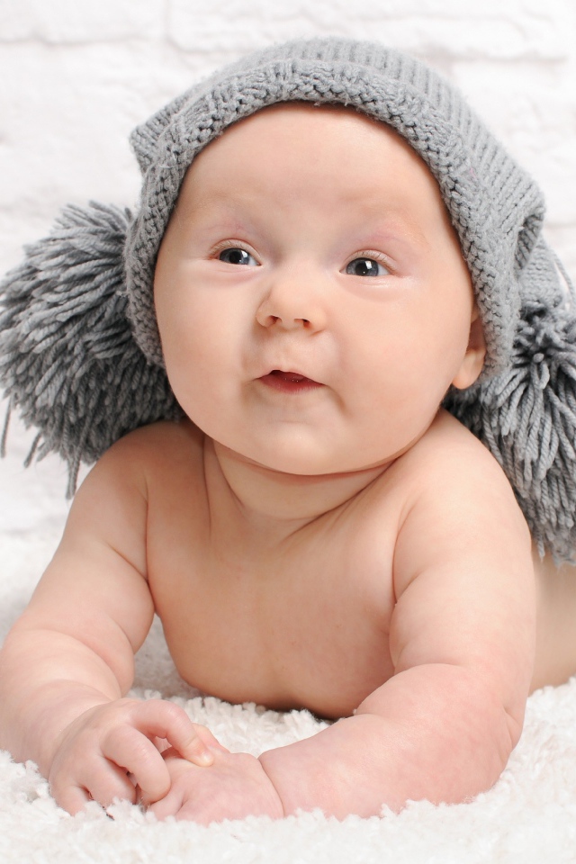 Улыбающийся грудной ребенок в серой шапке с бубонами