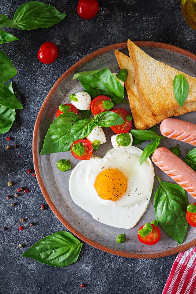 Яичница на тарелке с помидорами, гренками, сыром, сосисками и зеленью базилика