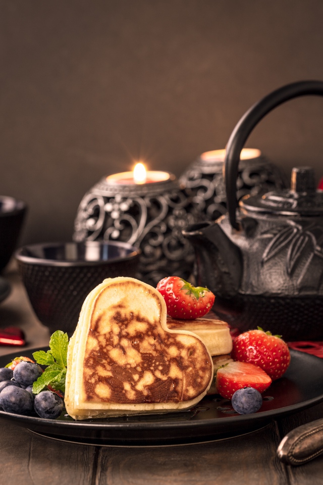 Оладьи в форме сердца с ягодами черники и клубники на столе с чаем и свечами