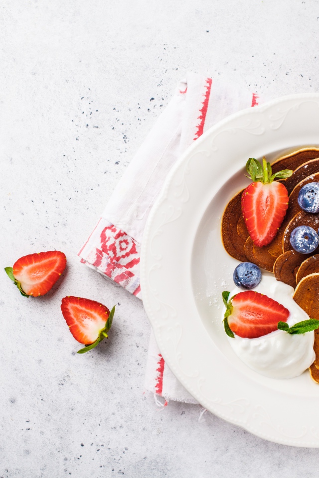 Оладьи со сметаной в тарелке с ягодами черники, клубники и красной смородины