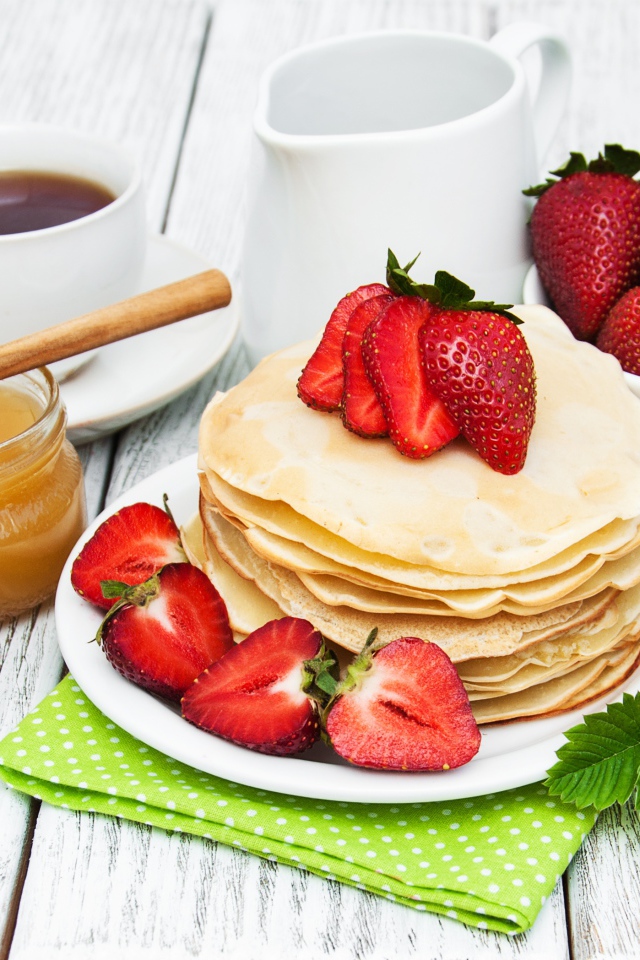 Тонкие блины на столе с ягодами клубники, медом, чаем и джемом