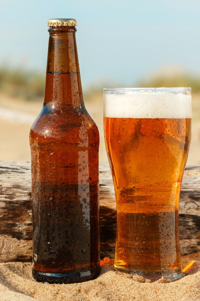 Бутылка пива и бокал стоят на песке 