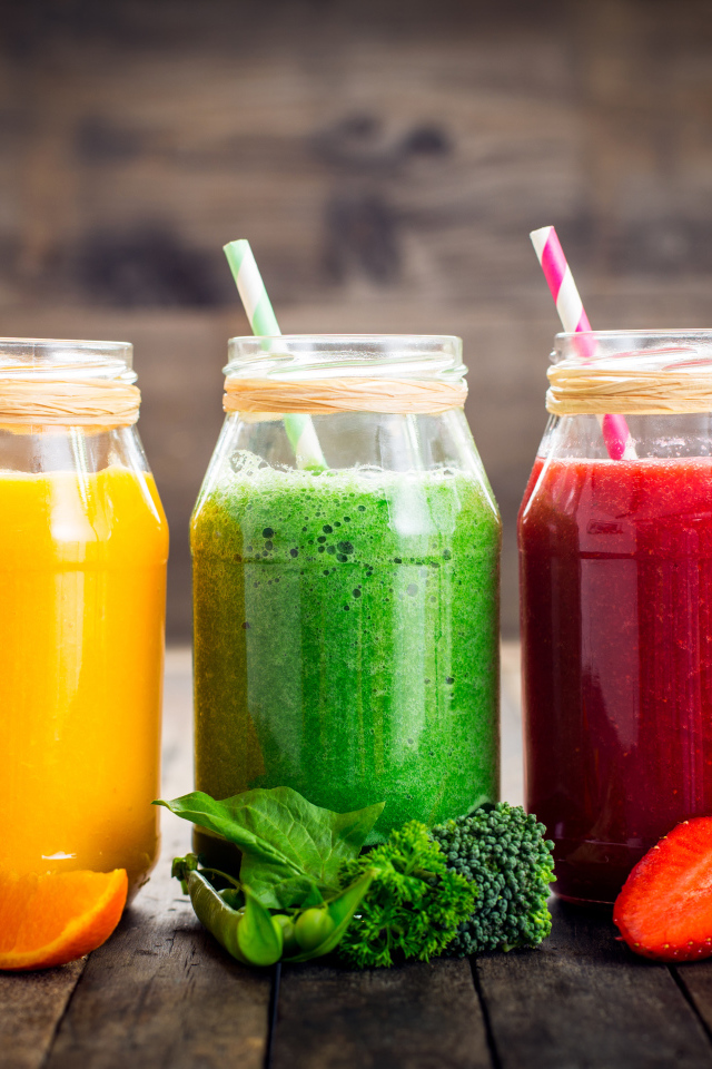 Три банки смузи с трубочкой на столе с апельсином, брокколи и клубникой