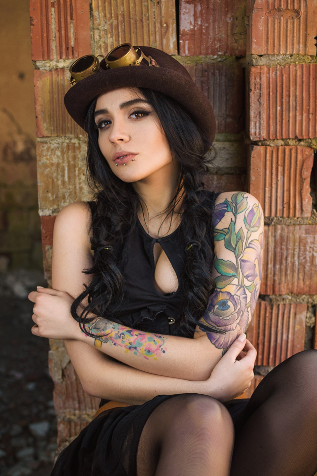 Девушка в шляпе с татуировками на руке сидит у стены