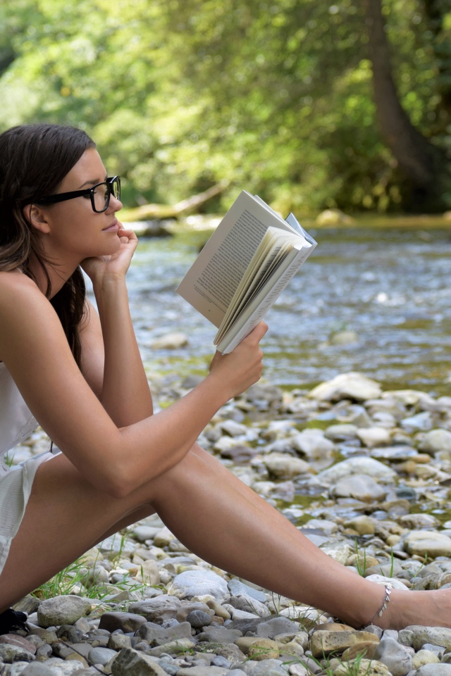 Молодая девушка в белом платье читает книгу на берегу реки