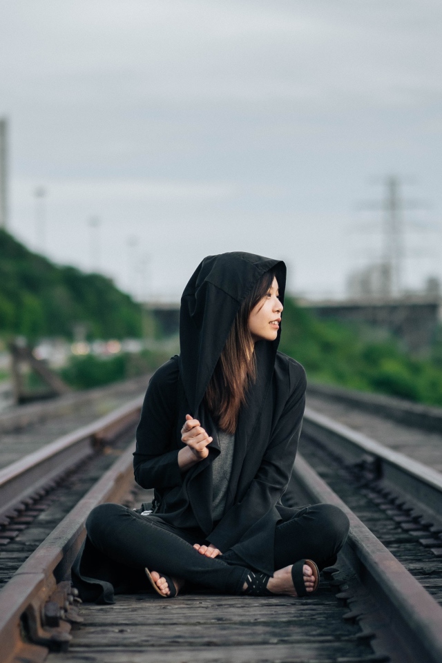 Девушка азиатка в черном балахоне сидит на рельсах 