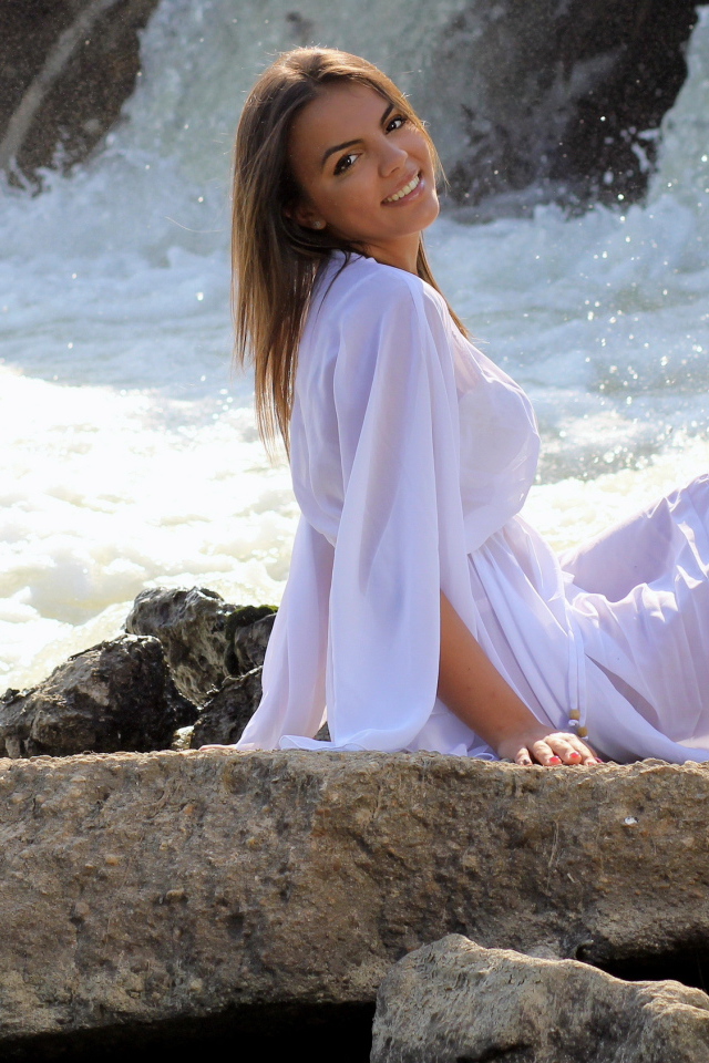 Красивая девушка в белом платье сидит на камне у воды 