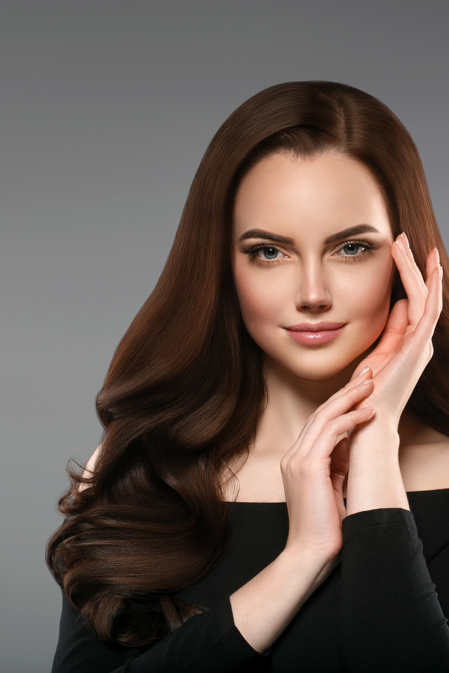 Красивая нежная длинноволосая шатенка  с руками у лица на сером фоне