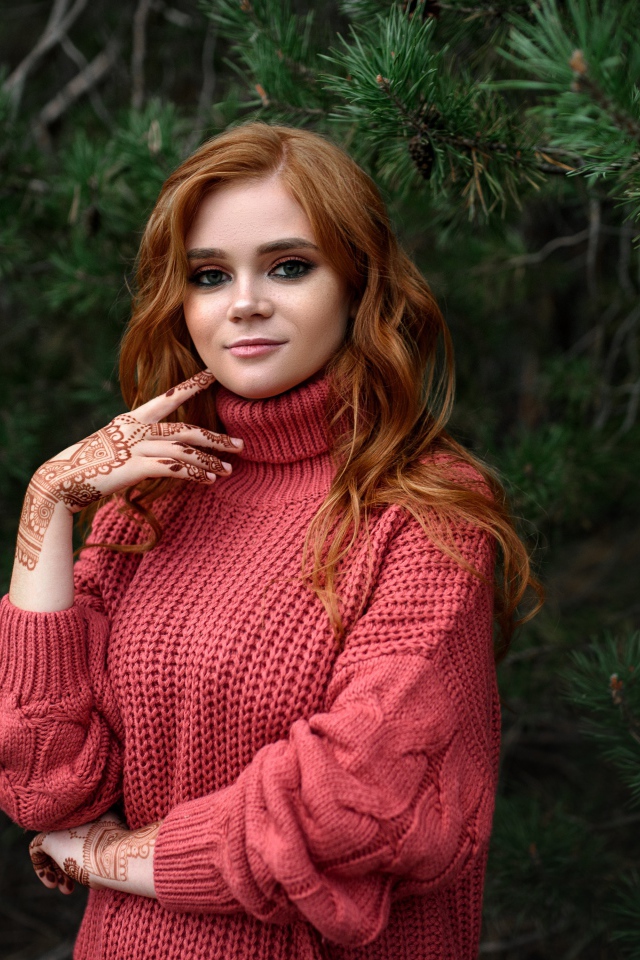 Рыжеволосая девушка в розовом свитере стоит у ели