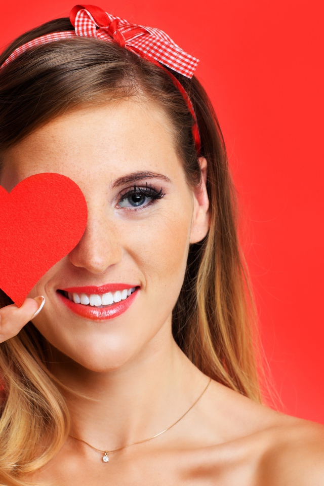 Улыбающаяся девушка с бумажным сердцем у лица на красном фоне