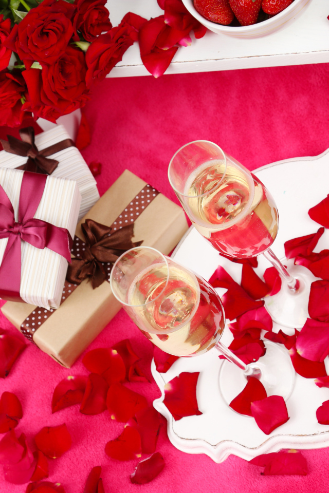 Два фужера с шампанским на красном покрывале с лепестками, подарками и букетом роз для любимой