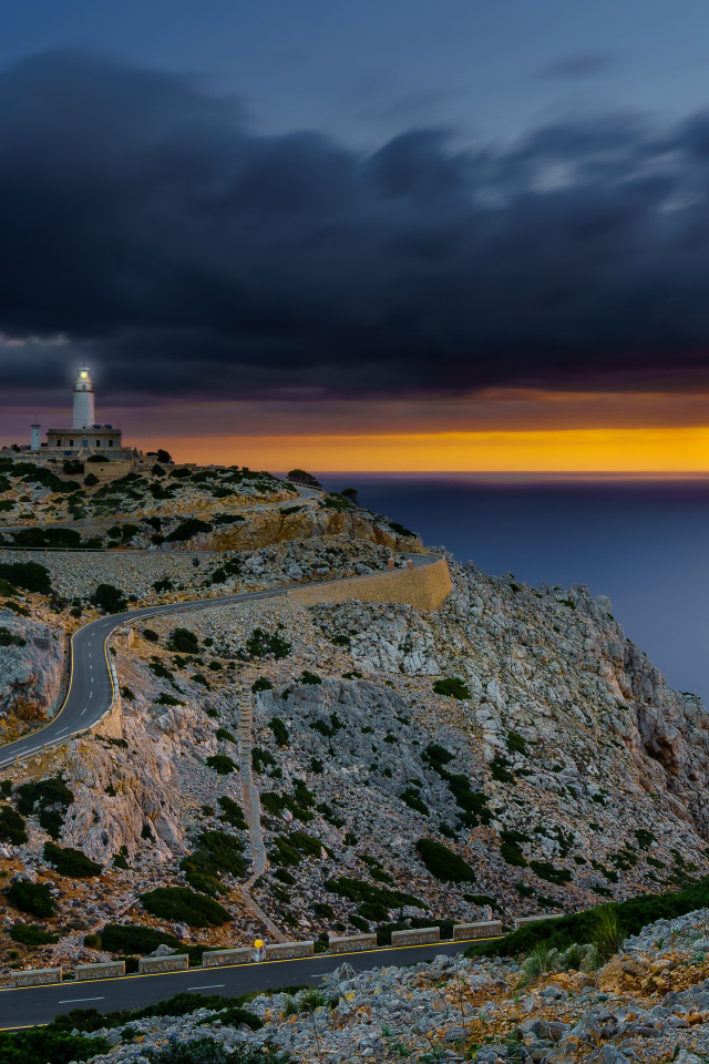 A winding road leads to a lighthouse on the edge of a cliff under a cloudy sky