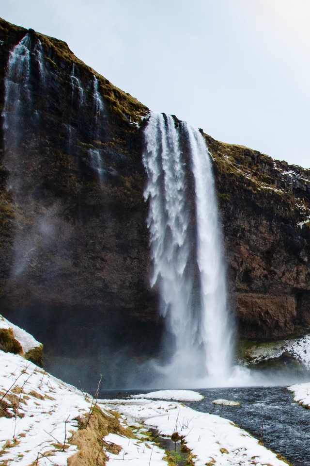 Водопад стекает со скалы в реку с покрытыми снегом берегами