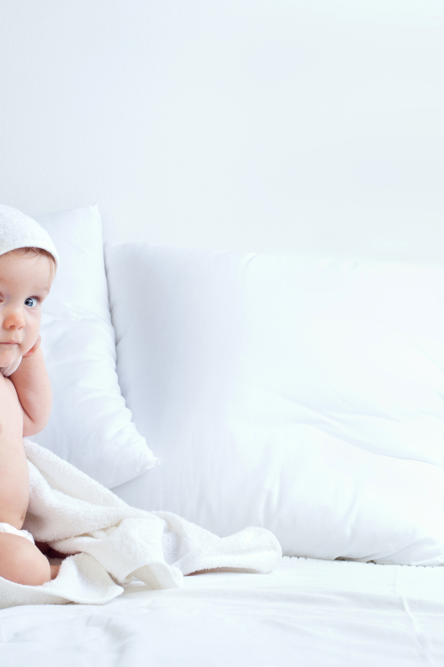 Маленький мальчик сидит в белом покрывале на кровати с подушками