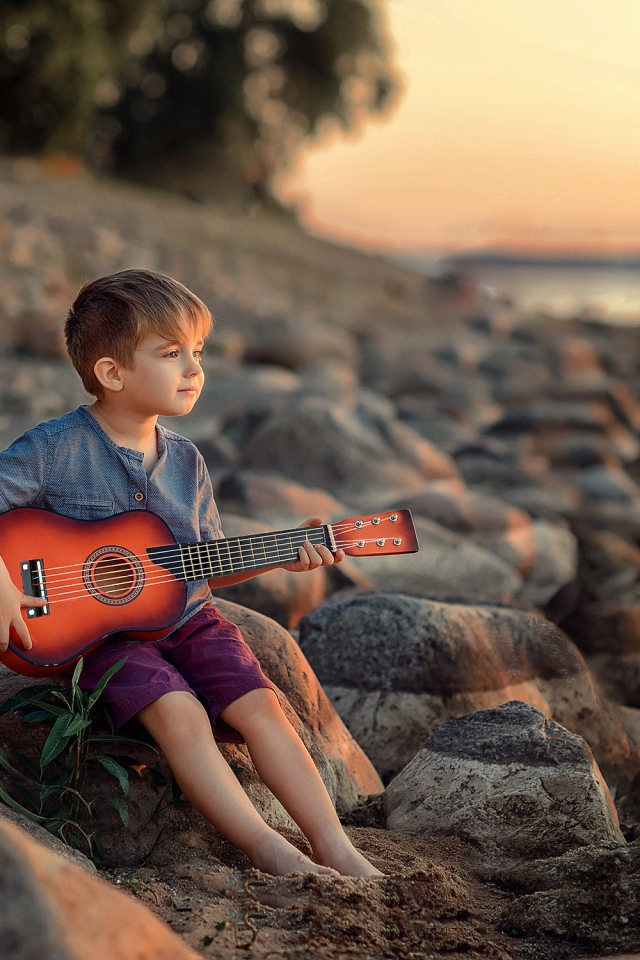 Маленький мальчик сидит с гитарой на камнях у моря 