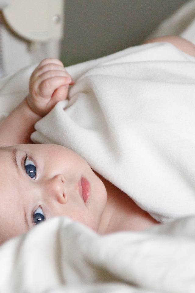 Маленький грудной голубоглазый ребенок лежит под белым покрывалом 
