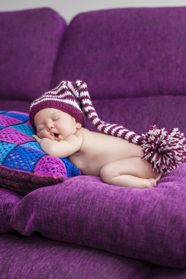 Маленький ребенок в большой шапке спит на фиолетовых подушках