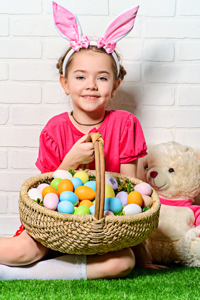 Улыбающаяся девочка сидит у стены с корзиной пасхальных яиц и игрушками
