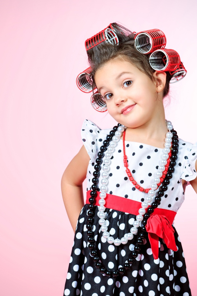 Маленькая девочка с бигудями на голове с феном в руке на розовом фоне