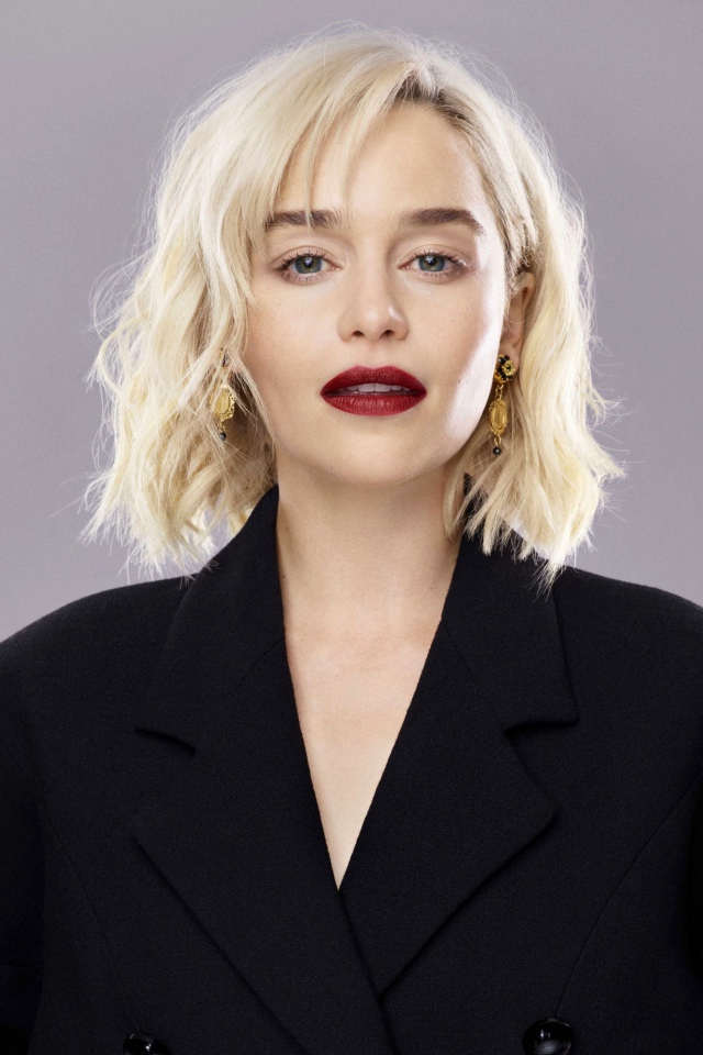 Яркая блондинка, актриса Эмилия Кларк в черном пальто на сером фоне