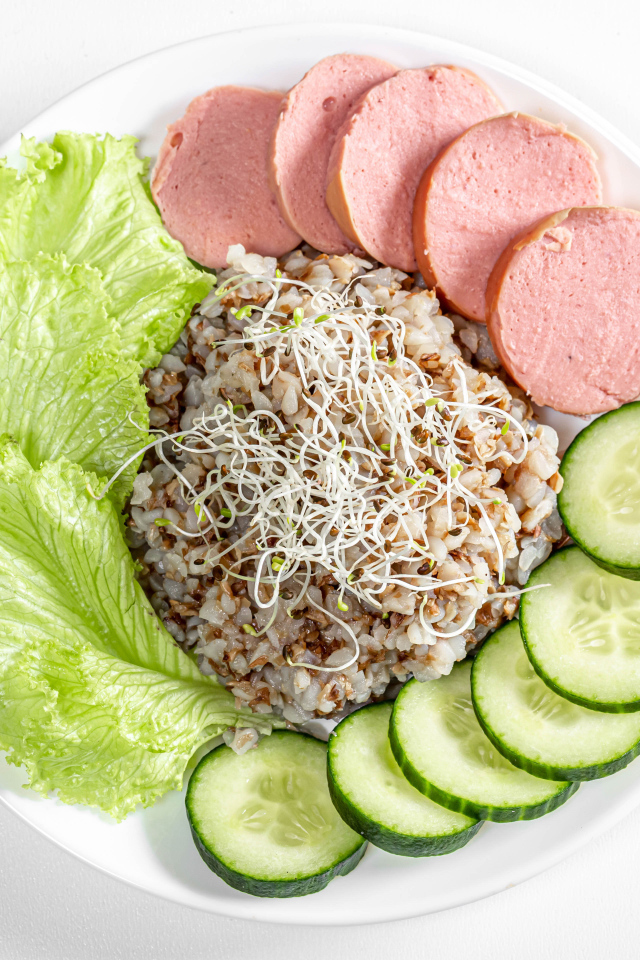 Гречка на тарелке с огурцом, колбасой и листьями салата на белом фоне