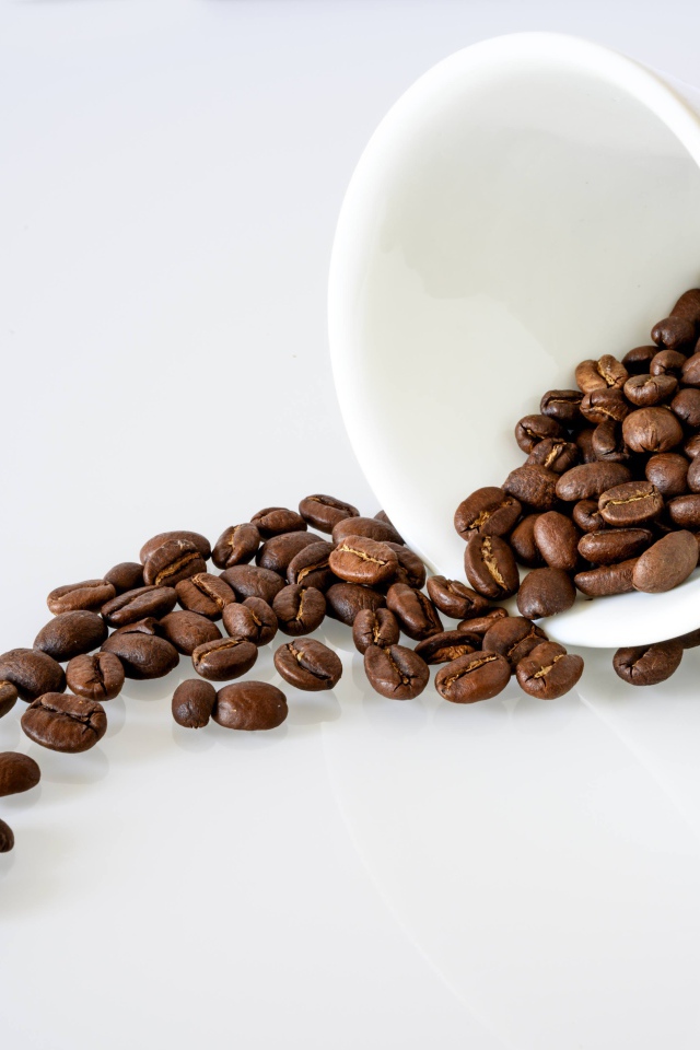 Кофейные зерна высыпают из белой чашки на стол 