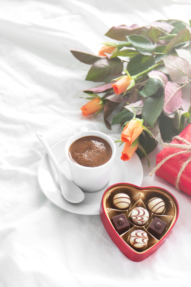 Чашка кофе на белой кровати с конфетами, подарком и букетом роз