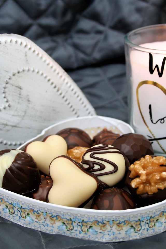 Шоколадные конфеты в коробке в форме сердца на кровати с зажженной свечой 