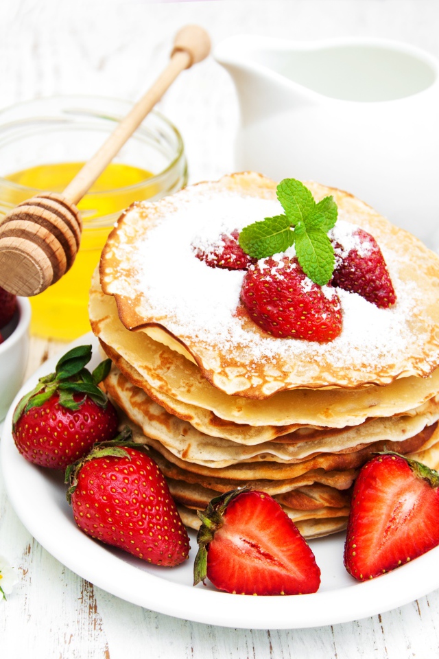 Тонкие блины с сахарной пудрой и ягодами клубники на столе с молоком и медом