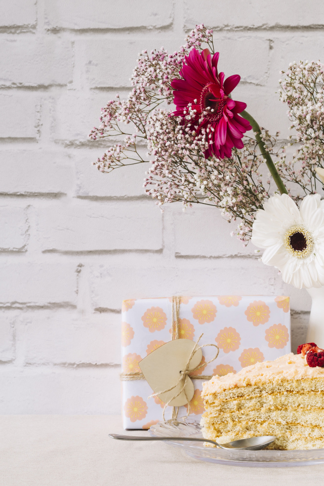 Кусок торта, подарок и букет на столе на фоне стены 