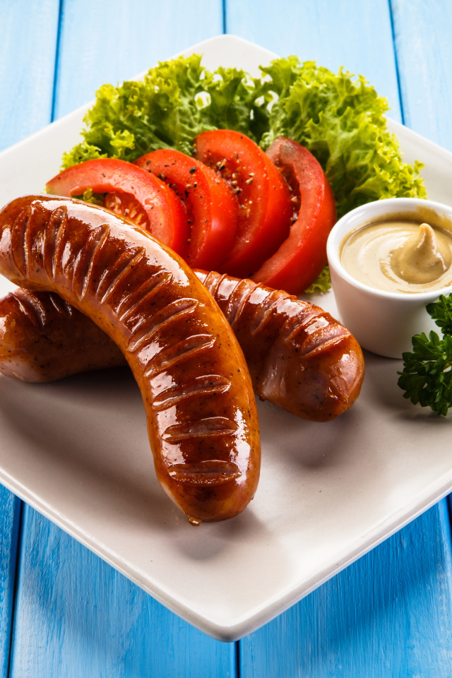 Две жареные колбаски на тарелке с помидорами, горчицей и листьями салата 