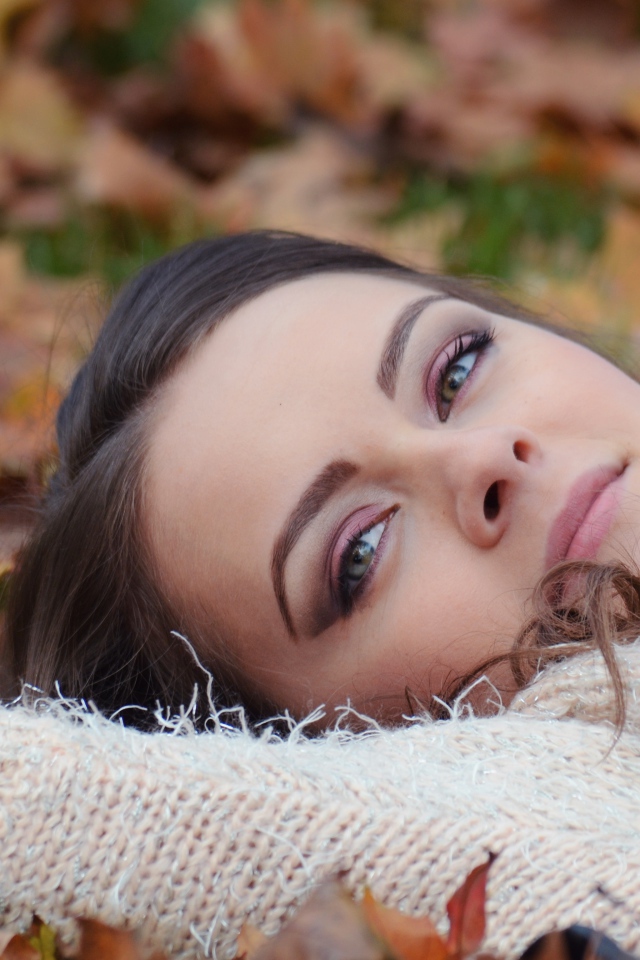 Красивая девушка в теплом свитере лежит на сухой листве 