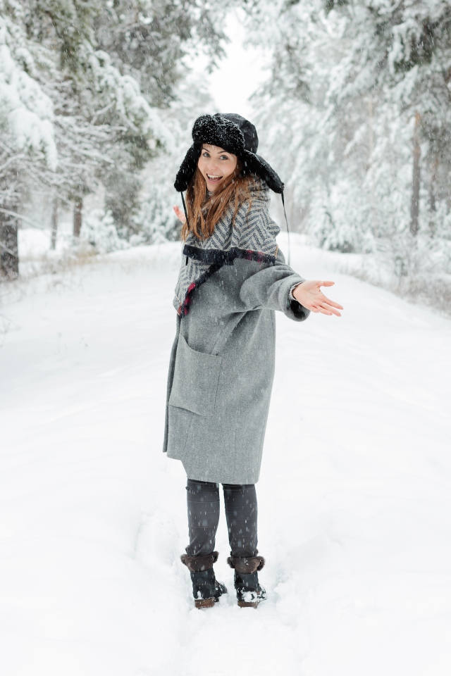 Девушка в пальто и теплой шапке гуляет в заснеженном лесу 