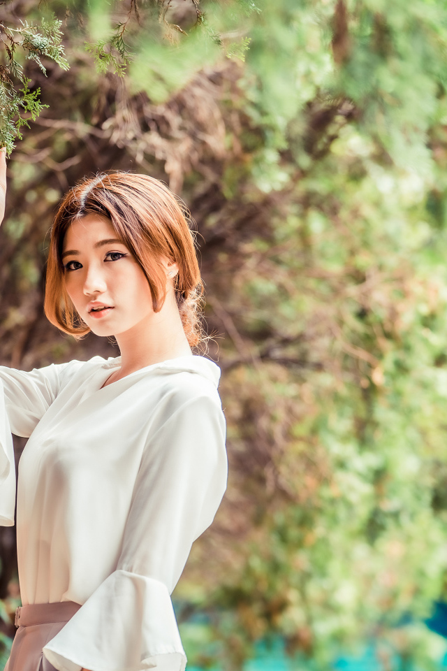 Молодая азиатка в белой блузке стоит у дерева 