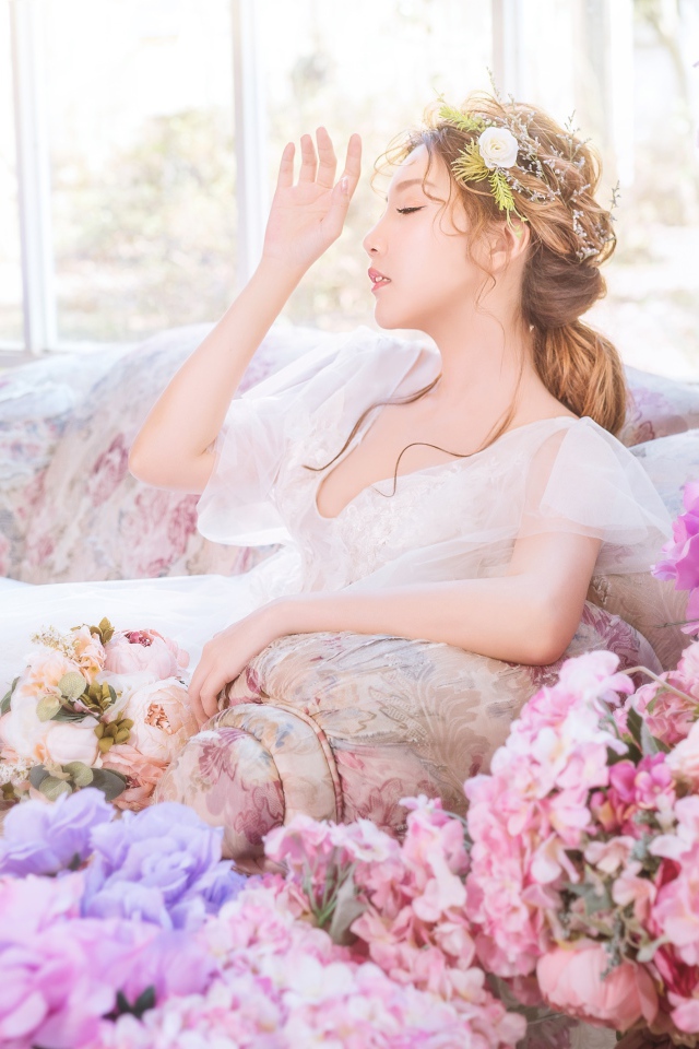Девушка азиатка в белом платье сидит на диване в комнате с цветами