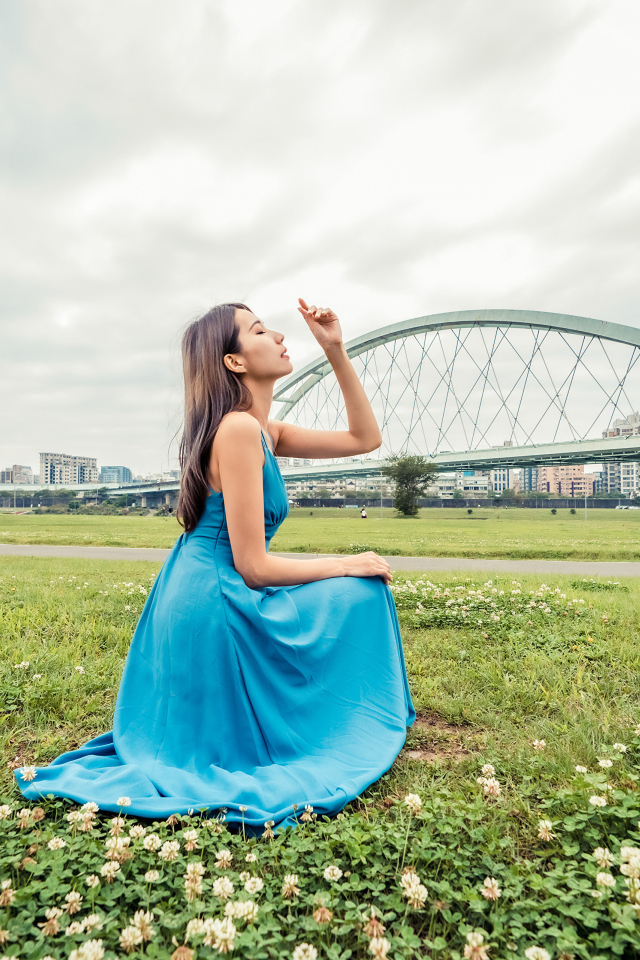 Красивая девушка азиатка в голубом платье  сидит на траве на фоне моста