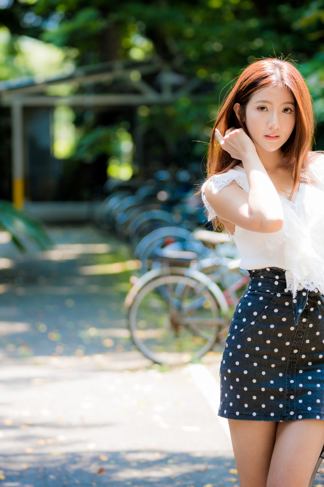 Красивая девушка азиатка в юбке в горох стоит у велосипеда 