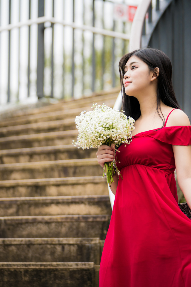 Красивая азиатка в красном платье стоит на ступеньках с букетом ландышей