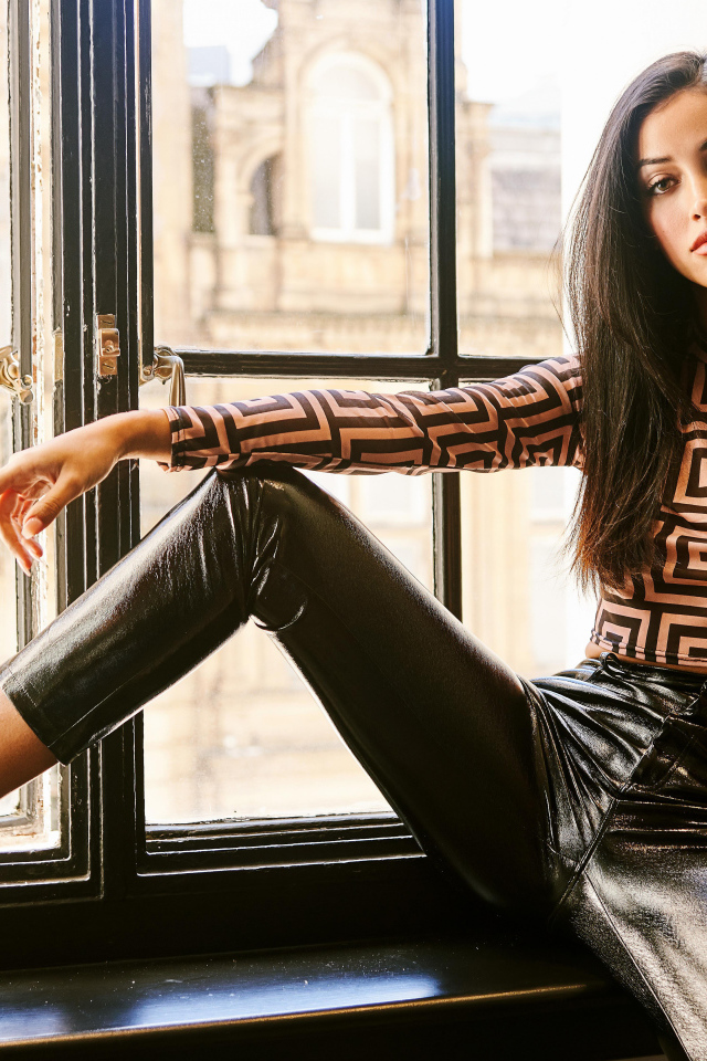 Красивая брюнетка в черных брюках сидит на окне 