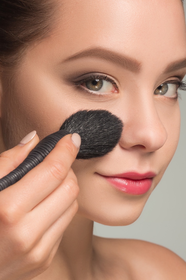 Красивой девушке наносят макияж на лицо кистью 