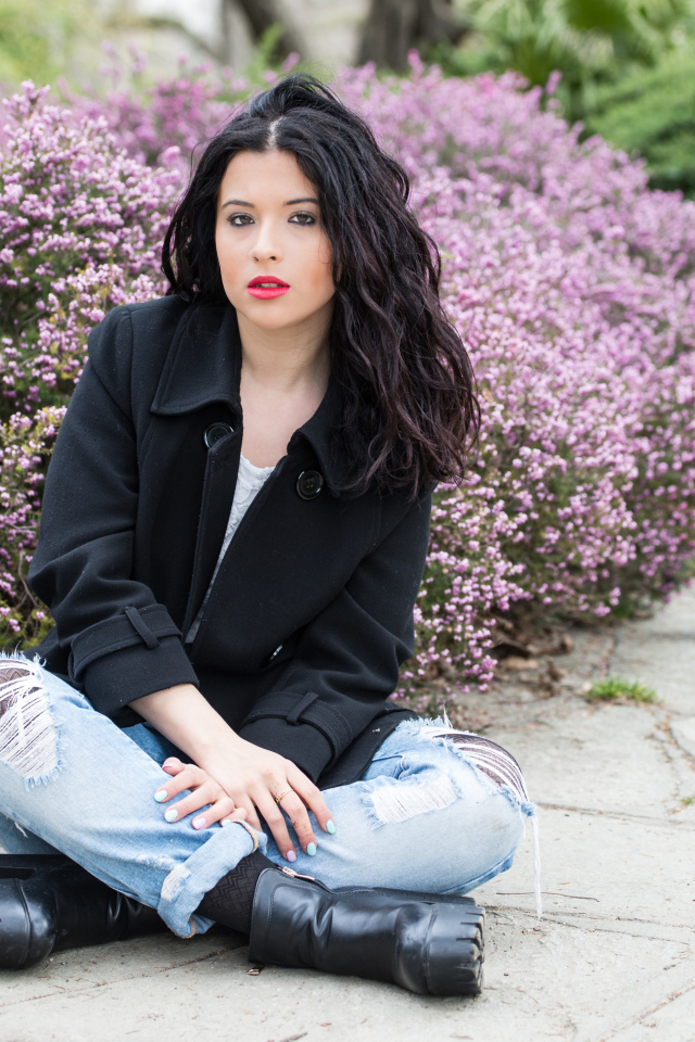 Красивая девушка в черном пальто сидит на земле у кустов