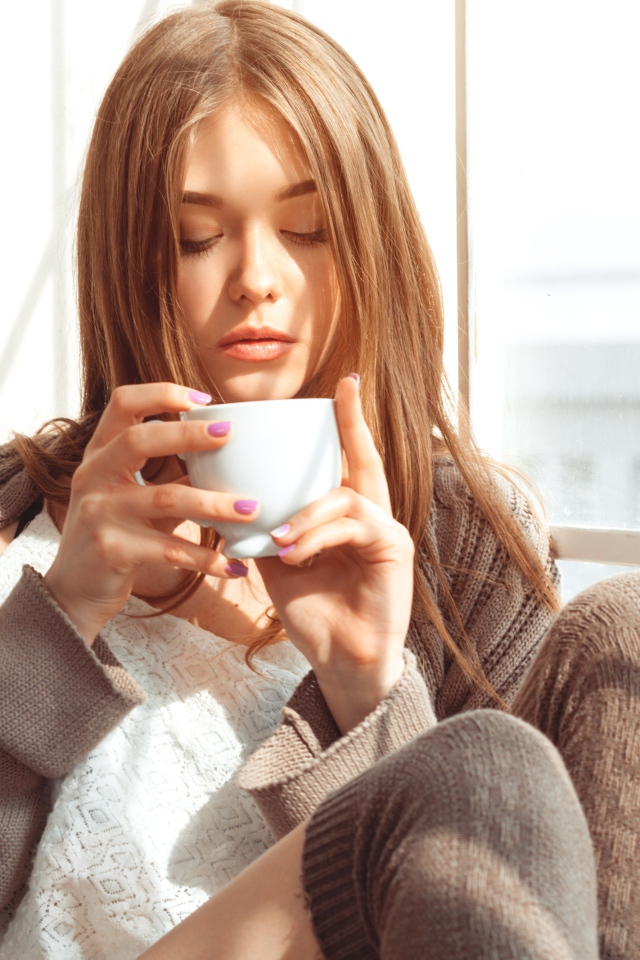 Красивая девушка в свитере пьет кофе у окна