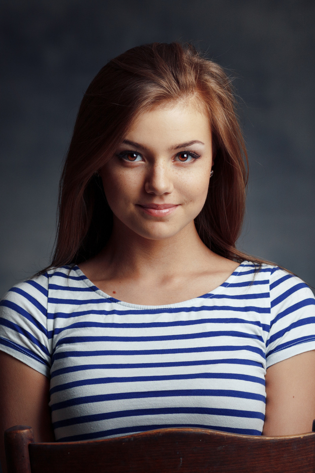 Милая кареглазая девушка в полосатой майке сидит на стуле 