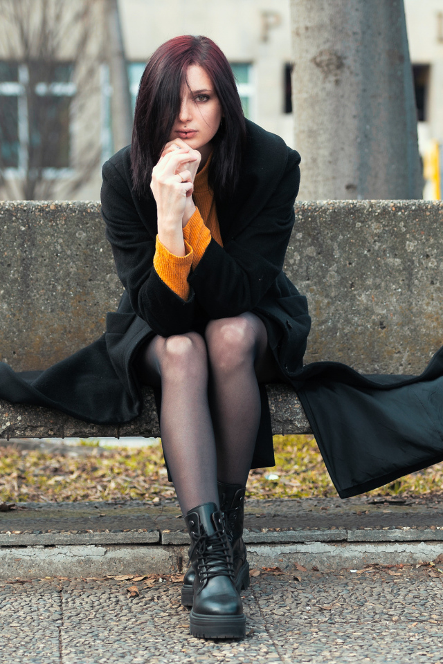 Девушка в черном пальто сидит на бетонной лавке 