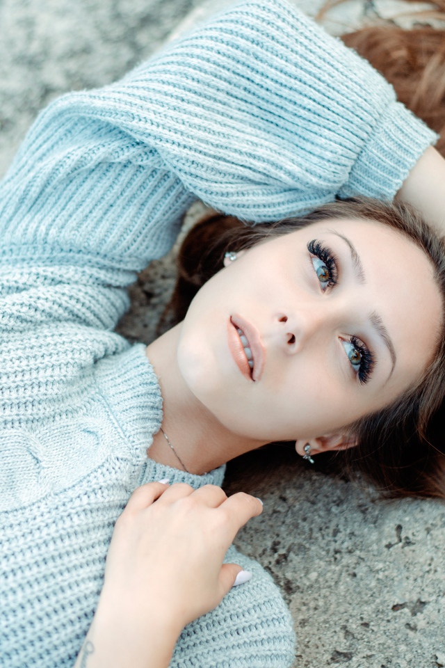 Длинноволосая девушка в голубом свитере лежит на земле 