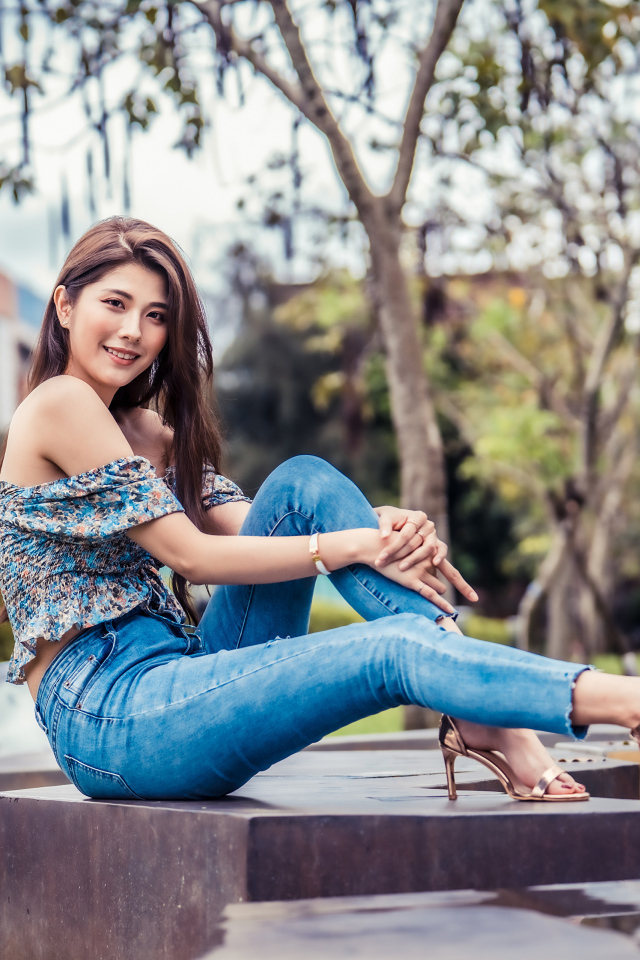Улыбающаяся азиатка в джинсах сидит в парке