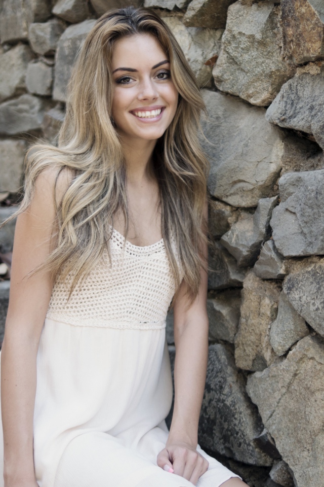 Улыбающаяся  девушка в белом платье стоит на ступеньках у стены