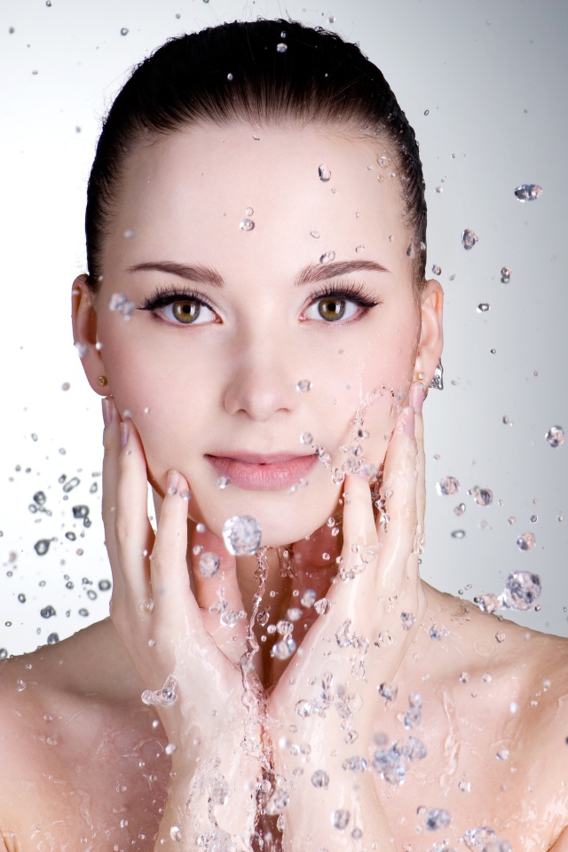 Брызги воды летят на красивое лицо молодой девушки на сером фоне