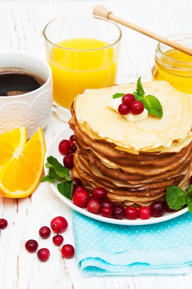 Тонкие румяные блинчики на столе с ягодами клюквы, апельсинами, медом и кофе