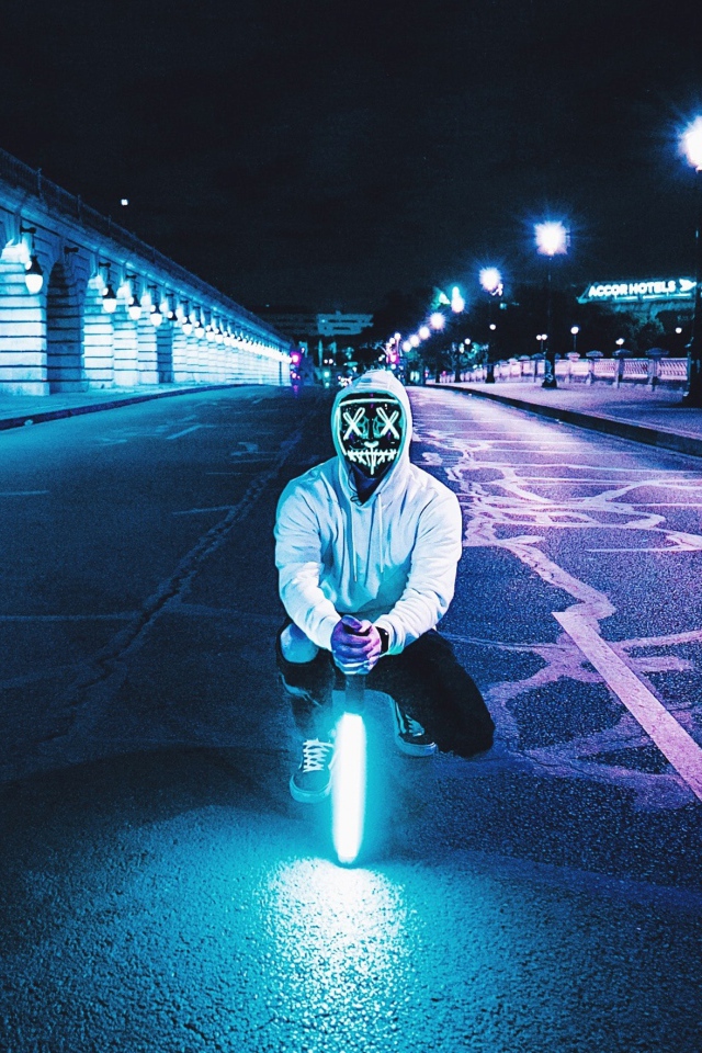 Парень в светящейся маске сидит с неоновой лампой на дороге